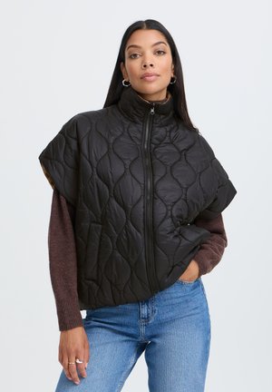 Femme aux longs cheveux foncés portant une veste sans manches matelassée noire par-dessus un pull marron et un jean bleu, debout devant un fond uni.