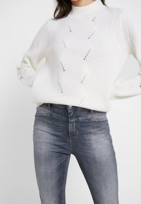 Suéter blanco de punto con un cuello alto, que presenta un patrón ondulado y puntos abiertos. Combinado con jeans gris ajustados.