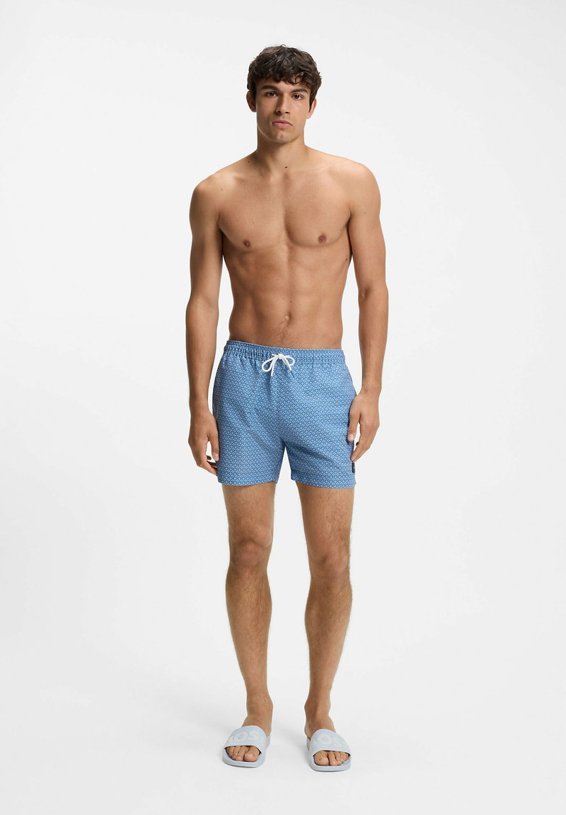 Jeune homme aux cheveux bouclés portant un short de bain bleu à motifs et des sandales bleu clair, debout devant un fond blanc uni.