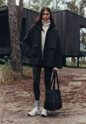 Une femme se tient sur un chemin forestier, à côté d’un bâtiment moderne et sombre. Elle porte un manteau noir matelassé et un cabas noir.