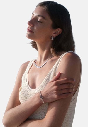 Woman with short brown hair in a sleeveless knit top, wearing silver necklace, hoop earrings, and a beaded bracelet, eyes closed, arms crossed.