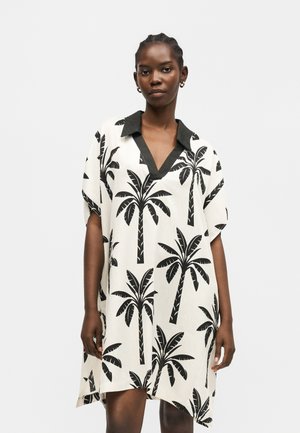 Woman wearing a loose white dress with black palm tree print and black collar, standing against a plain light background.