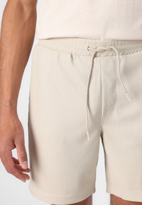 Pantaloni scurți bej din material cu nervuri, cu talie elastică și închidere cu șnur. Textură netedă, lungime până la mijlocul coapsei, fără modele sau grafice vizibile.