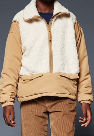 Osoba ubrana w beżowo-białą kurtkę z polaru sherpa z półzamkiem i przednimi kieszeniami z patkami, w połączeniu z brązowymi sztruksowymi spodniami.