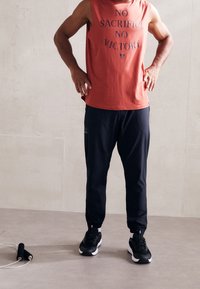 Rdzawy czerwonawy bezrękawnik z napisem, czarne sportowe spodnie oraz czarne sneakersy. Spodnie mają boczną kieszeń i zwężany krój.