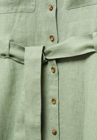 Robe en lin vert clair avec des boutons marron au centre et une ceinture en tissu nouée à la taille.