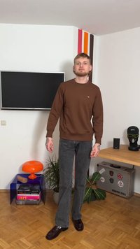 Brauner Sweatshirt, graue Schlagjeans und dunkle Schuhe. Der Sweatshirt hat ein kleines Logo; die Umgebung zeigt einen Fernseher, eine orange Lampe und eine Topfpflanze.