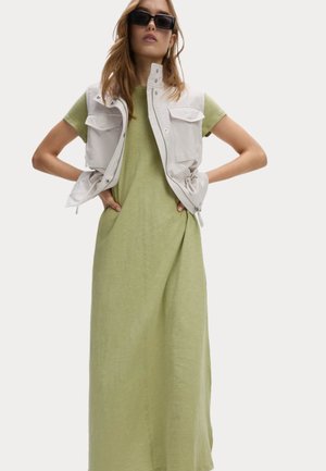 Lichtgroene linnen maxi-jurk gecombineerd met een witte vest met vier voorkwerpen. Model draagt zwarte zonnebril, met de armen op de heupen.