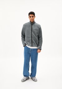 Giacca in pile grigia con colletto alto, chiusura con zip e tasca frontale, abbinata a jeans blu larghi e sneaker grigie. Design semplice, senza motivi.