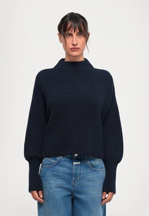 Woman with dark hair tied back wearing a navy blue ribbed sweater with balloon sleeves and high-waisted blue jeans against a white background.