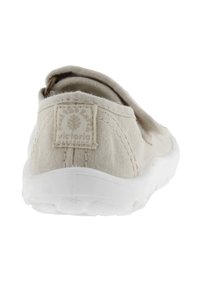 Zapato sin cordones de color beige claro con parte superior de lona, suela de goma blanca y una pequeña etiqueta de logo en la parte posterior. Presenta un diseño redondeado.