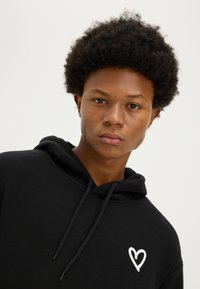 Young person with curly hair wearing a black hoodie featuring a white heart design on the chest, against a plain light background.