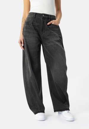 Person wearing loose blackwashed wide-leg jeans with raw hems and white sneakers, one hand in pocket, against a plain background.