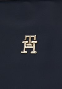 Navy kleurige stoffen tas met een opvallend gouden logo met hoekige lijnen en een gestructureerd oppervlak. Het materiaal heeft een subtiele glans.