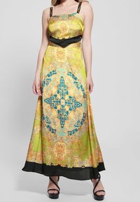 Lange, mouwloze jurk in een levendig groen met complexe gouden en blauwe patronen. Heeft een zwarte band in de taille en dunne bandjes. Soepele textuur.