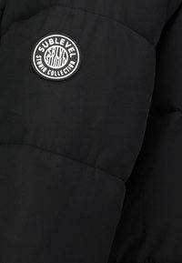 Schwarze Daunenjacke mit einem runden Logopatch und dem Schriftzug "Sublevel Studio Collection", die eine glatte Textur und ein gestepptes Design aufweist.