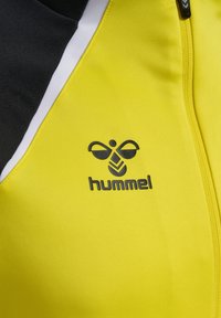 Chaqueta atlética amarilla con acentos en negro, que cuenta con cierre de cremallera y un logo impreso en negro. Tejido suave y ligero con un diseño deportivo.