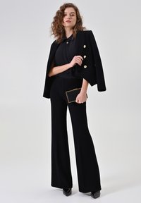 Zwart op maat gemaakt pak met een gestructureerd jasje met gouden knopen, wijde broekspijpen en een textuurrijke afwerking. Gecombineerd met een zwarte clutch.