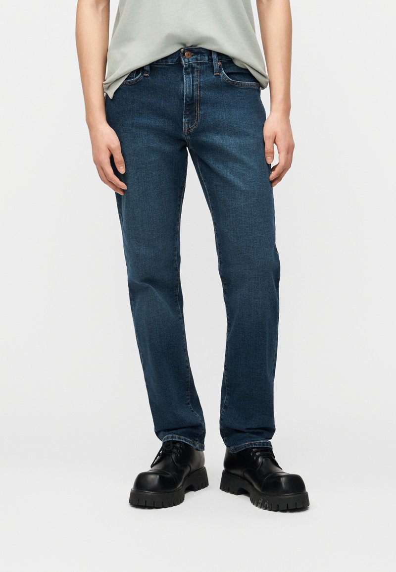 Άτομο που φοράει σκουρό μπλε τζιν με ίσια γραμμή, ανοιχτό γκρι μπλουζάκι μέσα από το παντελόνι και μαύρα παπούτσια με χοντρή σόλα και κορδόνια, στέκεται ακίνητο.