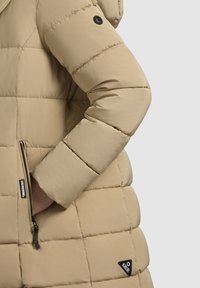 Beige pufferjakke med quiltet tekstur, der har en lynlåslomme, sorte hardware-detaljer og en hætte. Nærbillede af ærmet og siden.