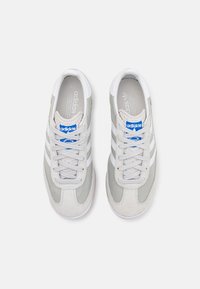 Grå och vita mockasinsandaler med tre vita ränder, runda snören och en blå logotags på tungan. Platt gummisula.