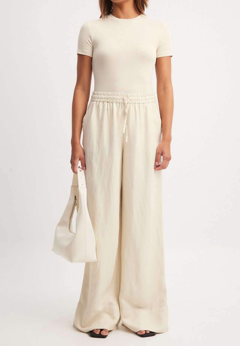 Beige aansluitend T-shirt gecombineerd met losse, lichtbeige wijde broek en een bijpassende handtas. Gladde textuur, neutrale kleurenpalet.
