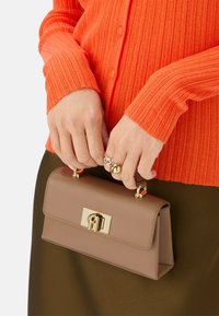 Borsa in pelle tan con hardware dorato, forma rettangolare, tenuta in mano. È visibile un maglione arancione a coste, che aggiunge texture e contrasto di colore.