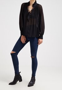 Blusa nera a pieghe con scollo a cravatta, abbinata a jeans skinny scuri strappati e stivali alla caviglia neri. Tessuto morbido e leggero.