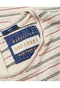 Vitt tyg med röda och svarta ränder, med en blå etikett med "SUPERDRY" präglat i guld, som visar storlek "Small" och varumärkesinformation.