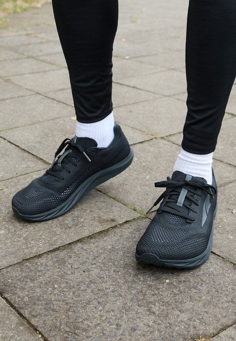 Black mesh sneakers with a textured upper, flat laces, and a cushioned sole. Worn with white socks and black leggings on a pavement surface.