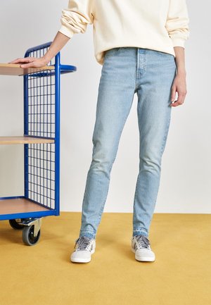 Personne portant un jean bleu clair et des baskets blanches, debout sur un sol jaune, reposant la main gauche sur une étagère métallique bleue avec des étagères en bois.