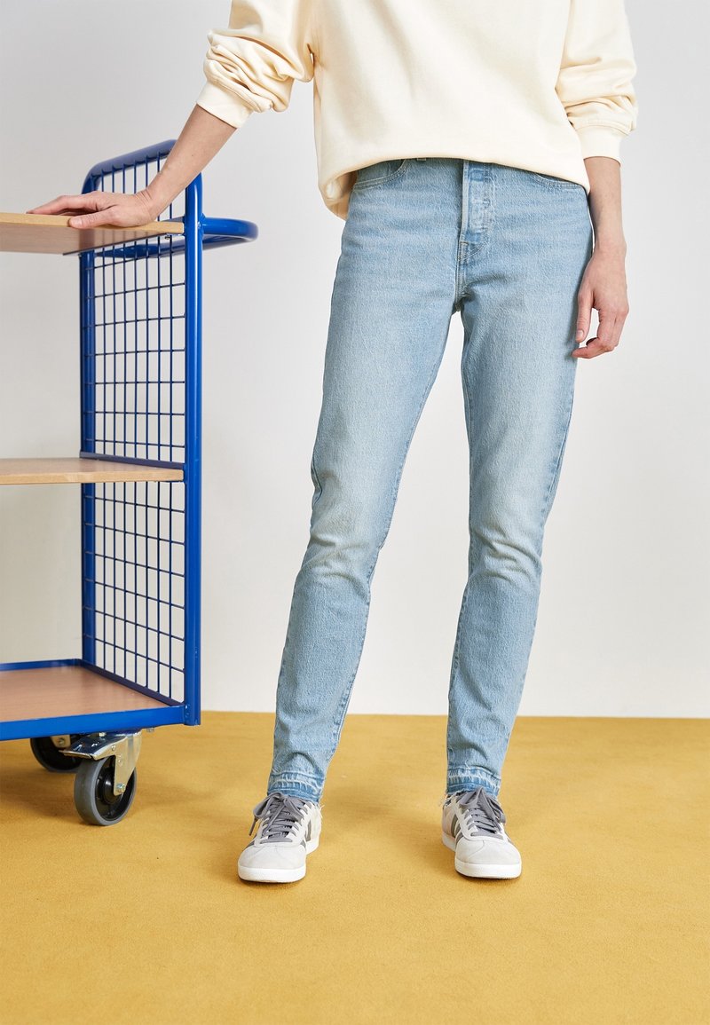 Personne portant un jean bleu clair et des baskets blanches, debout sur un sol jaune, reposant la main gauche sur une étagère métallique bleue avec des étagères en bois.