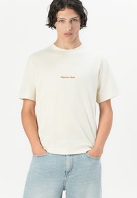 Krem-obilni pamučni T-shirt s kratkim rukavima, na prednjem dijelu s tekstom "Original Studio" u smeškastom vezu.