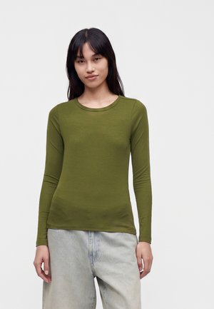 Jeune femme aux cheveux lisses et foncés, portant une chemise à manches longues vert olive et un jean ample gris clair, debout devant un fond uni.