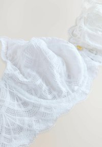 Reggiseno bianco in pizzo con intricati motivi geometrici e bordi ondulati. Tessuto morbido con un aspetto trasparente, caratterizzato da dettagli in metallo dorato.