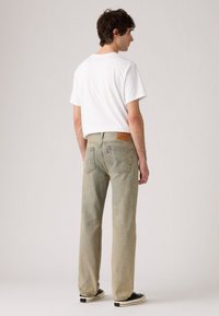 Hombre de espaldas que lleva una camiseta de manga corta blanca, jeans azul desgastados y zapatillas negras de perfil bajo, contra un fondo claro y sencillo.