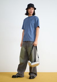 Tricou albastru cu manșon scurt și un logo subtil, asortat cu blugi gri deschis, largi, cu un gradient. Pălărie neagră tip bucket și geantă argintie de umăr.