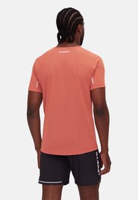 Man with braided hair wearing a coral short-sleeve shirt and black shorts with "Mammut" logos, facing away against a plain background.