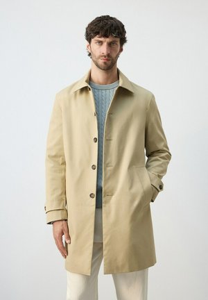 Mężczyzna w beżowym, jednorzędowym płaszczu przeciwdeszczowym, nałożonym na jasno niebieski sweter w warkocze i białe spodnie, stoi na jednolitym tle.