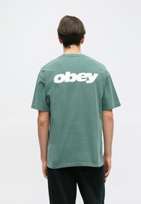 Roheline puuvillane T-särk, mille seljal on suur valge "obey" logo. Lühikesed varrukad ja lõtv istuvus, pehme tekstuur.