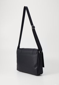 Sac à bandoulière noir en matériau lisse, de forme rectangulaire, avec une bandoulière ajustable et un subtil détail de couture sur le rabat.