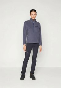 Ženska v temnem flis puloverju z žepi na zadrgo in črnimi športnimi hlačami, ki stoji proti enolični svetli ozadju.