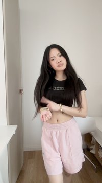 Νεαρή γυναίκα με μακριά μαύρα μαλλιά, που φοράει μαύρο κορμάκι και ροζ σορτς, δείχνοντας τα χρυσά βραχιόλια στον καρπό της μέσα σε εσωτερικό χώρο.