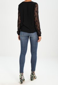 Schwarzes Oberteil mit langen Spitzensäumen, kombiniert mit hellblauen Jeans und gemusterten Ankle Boots, die florale und Camouflage-Designs zeigen.