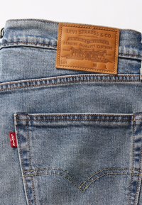 Jeans de mezclilla azul con un parche de cuero marrón que presenta texto en relieve. Etiqueta roja en el bolsillo trasero, con costuras doradas en contraste.