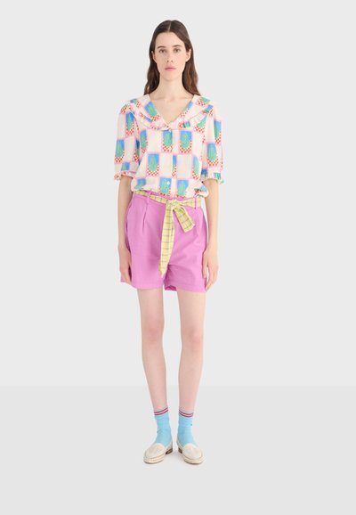 Femme portant un chemisier à motifs avec un col à volants, un short rose avec une ceinture à carreaux jaunes, des chaussettes bleu clair et des chaussures blanches, debout.