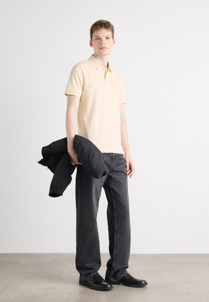 Beige Poloshirt, schwarze Steppjacke in der Hand, dunkelgraue Jeans und schwarze Schuhe, vor einem schlichten hellen Hintergrund.