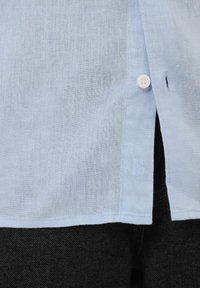 Ourlet de chemise en lin bleu clair avec un bouton blanc attaché au-dessus d'un jean en denim foncé.