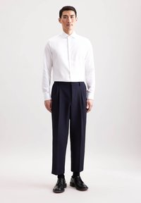 Camicia bianca a maniche lunghe con colletto alto, abbinata a pantaloni a pieghe blu navy e scarpe eleganti nere. Design semplice, texture liscia.