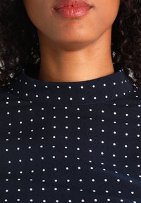 Parte inferiore del viso e collo di una donna che indossa una maglia blu navy con pois e colletto alto, con capelli scuri ricci strettamente arricciati.